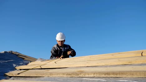 Adult-professional-builder-in-hardhat-measuring-length-of-wood-lumber-on-site-under-blue-sky,-slow-motion