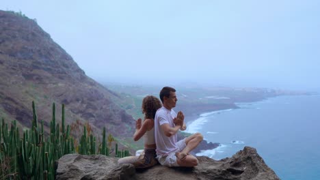 Man-and-woman-sitting-on-top-of-a-mountain-on-a-rock-back-to-back-meditate-and-do-yoga-on-the-background-of-the-ocean.