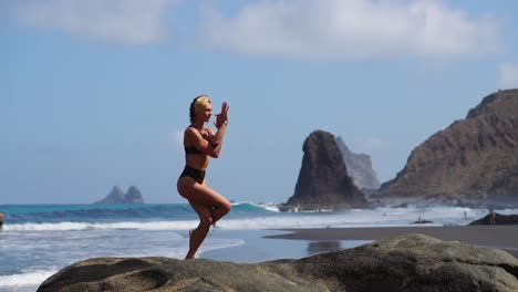 Junges-Mädchen-im-Bikini,-balancieren-auf-einem-Bein-tut-Yoga-steht-auf-einem-Felsen-am-Meer-auf-einem-schwarzen-Sandstrand