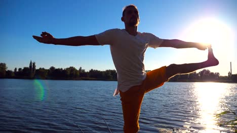 Young-man-standing-at-yoga-pose-on-wooden-jetty-at-lake.-Athlete-balancing-on-one-leg-at-nature.-Sporty-guy-doing-stretch-exercise-outdoor.-Concept-of-healthy-active-lifestyle.-Slow-motion-Close-up