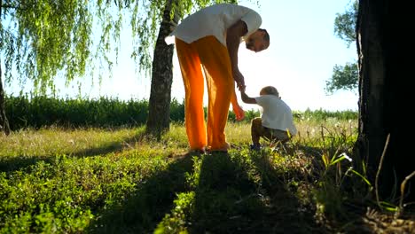 Cute-boy-finding-something-in-grass-while-walking-with-father-through-green-park-at-sunny-summer-day.-Young-dad-and-little-son-spending-time-together-outdoor.-Low-angle-view-Slow-motion-Close-up