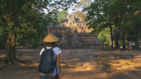 Slow-motion:-one-tourist-visiting-Angkor-ruins-at-sunrise,-travel-destination-Cambodia.-Woman-with-traditional-hat-walking-to-Baphuon-temple-in-the-jungle,-rear-view.