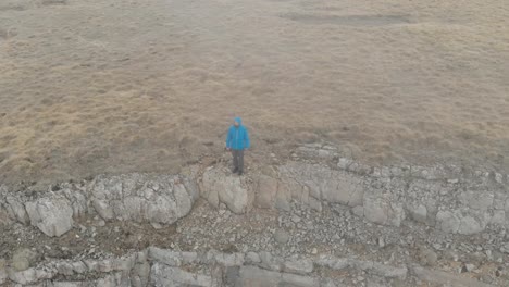 Hiker-man-in-a-blue-jacket-with-backpack-is-walking-and-standing-on-the-steep-edge-of-mountain-plateau-above-the-clouds.-Aerial-view.-Drone-is-orbiting