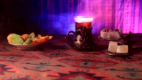 Arabian-tea-in-vintage-glass-with-eastern-snacks-on-a-carpet.-Eastern-tea-ceremony-on-dark-background-with-lights-and-smoke.-Empty-space.-Selective-focus.-Slider-shot