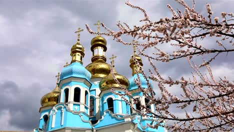 Orthodox-temple,-Clouds-above-the-temple,-golden-domes,-Timelapse,-exterior,-a-view-from-below,-View-through-the-branches-of-a-flowering-tree,-spring-landscape,-Blooming-tree