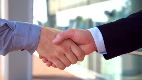 Close-up-of-two-successful-businessmen-greeting-each-other-in-urban-environment.-Young-colleagues-meeting-and-shaking-hands-near-office-building.-Handshake-of-business-partners-outdoor.-Side-view