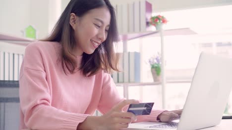 Beautiful-Asian-woman-using-computer-or-laptop-buying-online-shopping-by-credit-card-while-wear-sweater-sitting-on-desk-in-living-room-at-home.-Lifestyle-woman-at-home-concept.