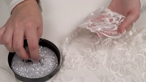 A-close-up-of-woman's-hands-holding-patterned-fabric-and-sewing-down-white-beads-from-the-lid-lying-in-front-of-her-to-embellish-it