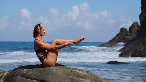Ein-junges-Mädchen-in-einem-Bikini,-balancieren-auf-einem-Stein-sitzend-und-Übungen-für-die-Bauchmuskeln,-Beine-abwechselnd-anheben.-Training-auf-dem-Hintergrund-des-Ozeans-auf-den-schwarzen-Sandstrand