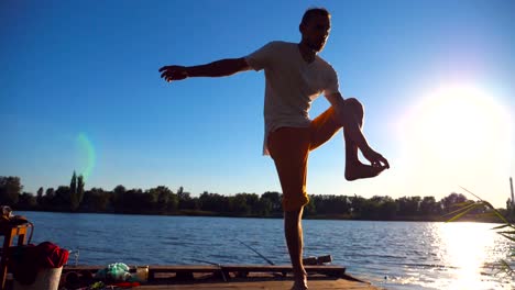 Young-man-standing-at-yoga-pose-on-wooden-jetty-at-lake.-Athlete-balancing-on-one-leg-at-nature.-Sporty-guy-doing-stretch-exercise-outdoor.-Concept-of-healthy-active-lifestyle.-Slow-motion-Close-up