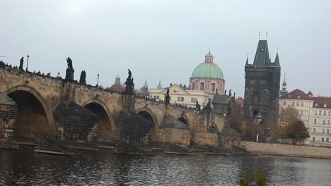 The-Charles-Bridge-on-the-background-of-the-old-tower-and-the-green-dome-of-the-cathedral-in-Prague,-side-view,-tourists-stroll-along-the-Charles-Bridge,-Prague,-October-19,-2017