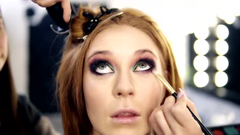 Front-footage-of-a-blonde-beautiful-model-sitting-and-looking-up-while-make-up-artist-finishing-her-smoky-eyes-.-Hairdresser-behind