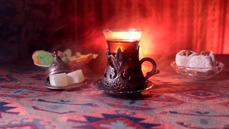 Arabian-tea-in-vintage-glass-with-eastern-snacks-on-a-carpet.-Eastern-tea-ceremony-on-dark-background-with-lights-and-smoke.-Empty-space.-Selective-focus.-Slider-shot