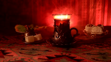 Arabian-tea-in-vintage-glass-with-eastern-snacks-on-a-carpet.-Eastern-tea-ceremony-on-dark-background-with-lights-and-smoke.-Empty-space.-Selective-focus.-Slider-shot