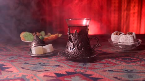 Arabian-tea-in-vintage-glass-with-eastern-snacks-on-a-carpet.-Eastern-tea-ceremony-on-dark-background-with-lights-and-smoke.-Empty-space.-Selective-focus.-Slider-shot