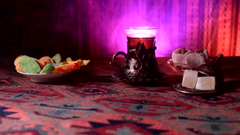 Arabian-tea-in-vintage-glass-with-eastern-snacks-on-a-carpet.-Eastern-tea-ceremony-on-dark-background-with-lights-and-smoke.-Empty-space.-Selective-focus.-Slider-shot