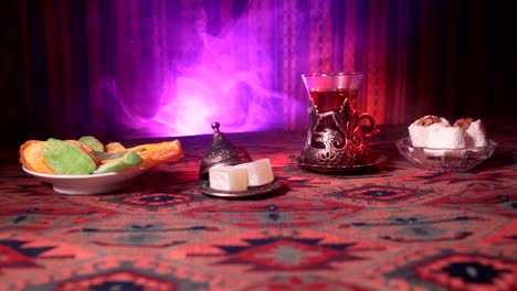 Arabian-tea-in-vintage-glass-with-eastern-snacks-on-a-carpet.-Eastern-tea-ceremony-on-dark-background-with-lights-and-smoke.-Empty-space.-Selective-focus.-Slider-shot