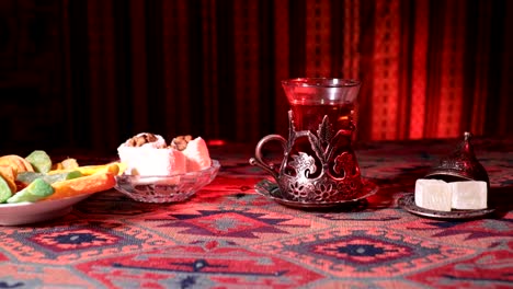 Arabian-tea-in-vintage-glass-with-eastern-snacks-on-a-carpet.-Eastern-tea-ceremony-on-dark-background-with-lights-and-smoke.-Empty-space.-Selective-focus.-Slider-shot