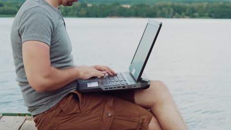 vielbeschäftigter-Mann-in-einem-grauen-T-shirt-sitzt-auf-einem-Mauerwerk-und-Texteingabe-hinter-einen-Laptop-auf-dem-Hintergrund-der-Natur