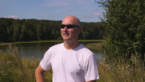 Portrait-adult-man-in-sunglasses-on-background-green-forest-and-lake-water