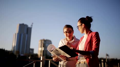 Dos-mujeres-con-los-planos-y-documentación-técnica-en-el-paseo-marítimo-en-el-fondo-de-los-rascacielos-en-construcción-para-discutir-los-riesgos-y-la-rentabilidad-de-la-construcción-de-un-nuevo-centro-de-negocios.