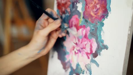 Close-up-shot-of-dirty-female-hand-painting-beautiful-bright-flowers-on-canvas-using-brush-and-paints.-Pictorial-arts,-creativity-and-artistry-concept.