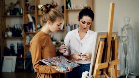 Freundlichen-Kunst-Lehrer-gut-aussehende-Frau-in-Freizeitkleidung-ist-Studentin-sprechen-dann-geben-ihre-Pinsel-Lehre,-Mädchen-lächelt-und-Mischen-von-Farben-auf-Palette.