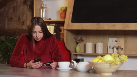 Young-woman-making-purchase-online,-typing-credit-card-details-on-black-smartphone-in-the-modern-kitchen