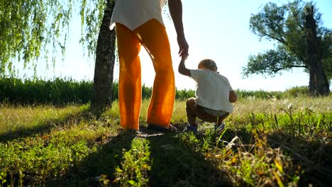 Cute-boy-finding-something-in-grass-while-walking-with-father-through-green-park-at-sunny-summer-day.-Young-dad-and-little-son-spending-time-together-outdoor.-Low-angle-view-Slow-motion-Close-up