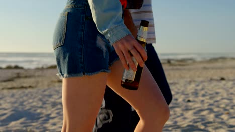 Side-view-of-couple-walking-with-beer-bottle-at-beach-on-a-sunny-day-4k