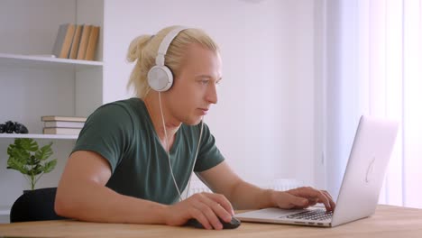 Nahaufnahme-Porträt-von-jungen-stattlichen-blonden-Hipster-Geschäftsmann-in-Kopfhörern-mit-Laptop-hören-Musik-freudig-drinnen-im-Büro