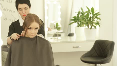 Hairdresser-wearing-cloack-robe-on-young-woman-before-haircut-in-hair-studio.-Preparing-for-haircut-in-hairdressing-salon-front-mirror