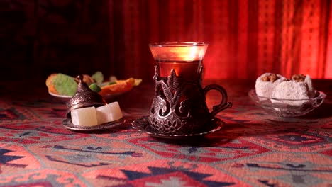 Arabian-tea-in-vintage-glass-with-eastern-snacks-on-a-carpet.-Eastern-tea-ceremony-on-dark-background-with-lights-and-smoke.-Empty-space.-Selective-focus.-Slider-shot