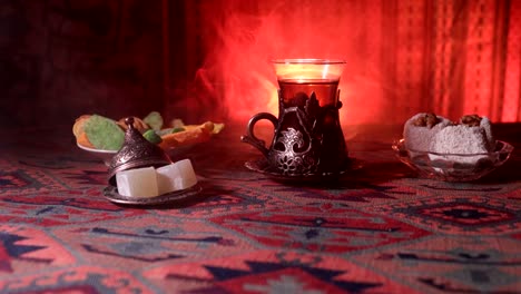 Arabian-tea-in-vintage-glass-with-eastern-snacks-on-a-carpet.-Eastern-tea-ceremony-on-dark-background-with-lights-and-smoke.-Empty-space.-Selective-focus.-Slider-shot