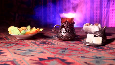 Arabian-tea-in-vintage-glass-with-eastern-snacks-on-a-carpet.-Eastern-tea-ceremony-on-dark-background-with-lights-and-smoke.-Empty-space.-Selective-focus.-Slider-shot