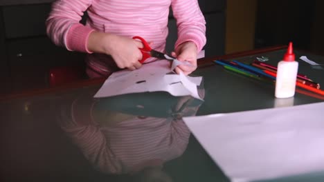 Close-up-shot-of-cute-little-girl's-hands-in-pink-sweater-cutting-shapes-from-paper,-glass-table-reflecting-her-face