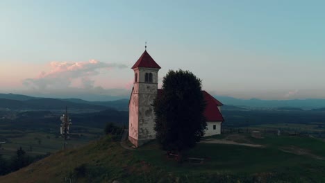 Fliegen-um-die-alte-katholische-Kirche-auf-einem-Hügel-mit-Aussicht-auf-das-Dorf-im-Sommer-in-den-Sonnenuntergang