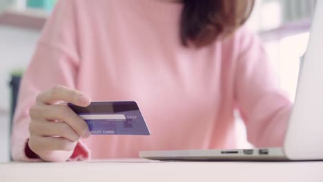 Beautiful-Asian-woman-using-computer-or-laptop-buying-online-shopping-by-credit-card-while-wear-sweater-sitting-on-desk-in-living-room-at-home.-Lifestyle-woman-at-home-concept.