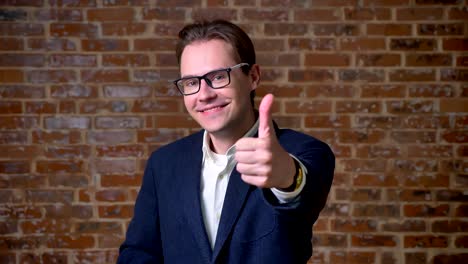 Cute-spectacled-caucasian-man-is-showing-like-sign-at-camera-and-smiling-sincerely-on-brick-background