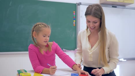 Individual-education,-Experienced-tutor-teaching-smart-girl-at-table-near-blackboard-in-classroom-of-School