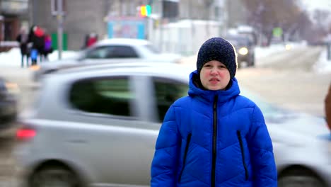 Boy-teenager-in-a-blue-down-jacket-standing-on-the-street.-Cars-are-riding-in-the-background,-the-boy-looks-around.