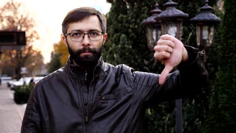 Retrato-de-joven-atractiva-barba-en-gafas-y-mirando-a-la-cámara.-Hombre-con-cara-de-poker-haciendo-pulgares-abajo,-lenta