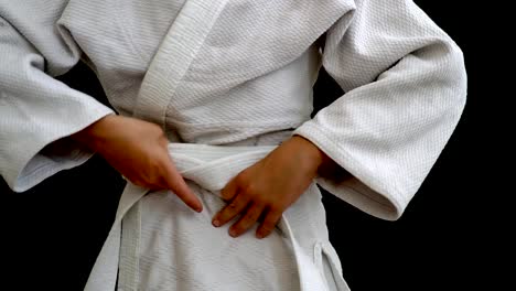 A-young-athlete-in-a-white-kimono-stands-on-a-dark-background.-The-young-man-holds-a-white-belt-in-his-hands,-his-hands-are-visible,-tying-the-belt-around-his-waist.-The-face-and-legs-are-not-visible