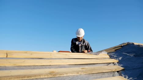 Adult-professional-builder-in-hardhat-measuring-length-of-wood-lumber-on-site-under-blue-sky