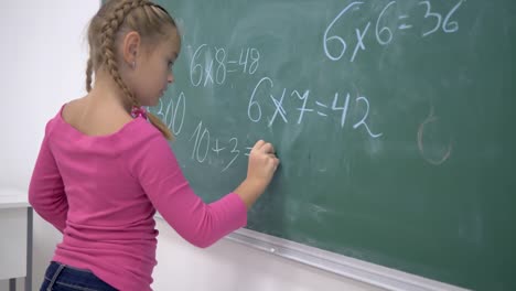 primary-education,-schoolgirl-with-chalk-solves-examples-on-mathematics-on-school-green-blackboard-in-class