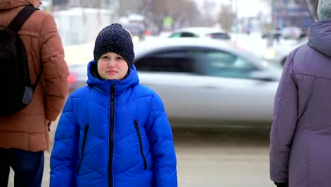 Boy-teenager-in-a-blue-down-jacket-standing-on-the-street.-Cars-are-riding-in-the-background,-the-boy-is-watching.