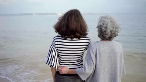 Rear-view-of-happy-senior-woman-embracing-tall-brunette-on-seashore.