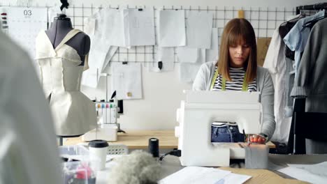 Self-employed-clothing-designer-is-working-with-sewing-machine,-stitching-textile-and-looking-at-sketches-lying-on-studio-table.-Everyday-work-at-tailor's-shop-concept.