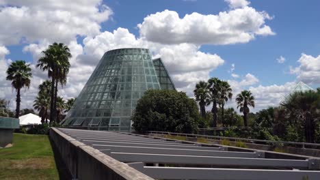 Panning-Video-of-Cone-Shape-Glass-Greenhouse-Surrounded-By-Palm-Trees-and-Blue-Sky