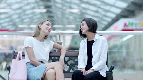 Fade-in-of-two-talkative-young-women-sitting-on-bench-near-railing-in-mall-and-chatting-after-shopping